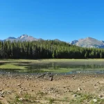 A panoramic view of a tranquil lake surrounded by trees and mountains on a clear day.